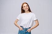 Cheerful young woman in T-shirt and denim pants holds hands in pockets on isolated white background.