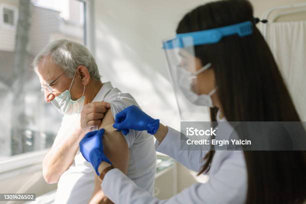 Female Doctor Wearing Protective Suit Injecting Vaccine Into Senior Patients Arm Stock Photo - Download Image Now