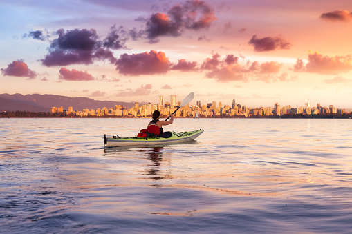 Beautiful woman sea kayaking in the ocean during a colorful and vibrant sunset with the city skyline in the background. Taken in Jericho, Vancouver, British Columbia, Canada.