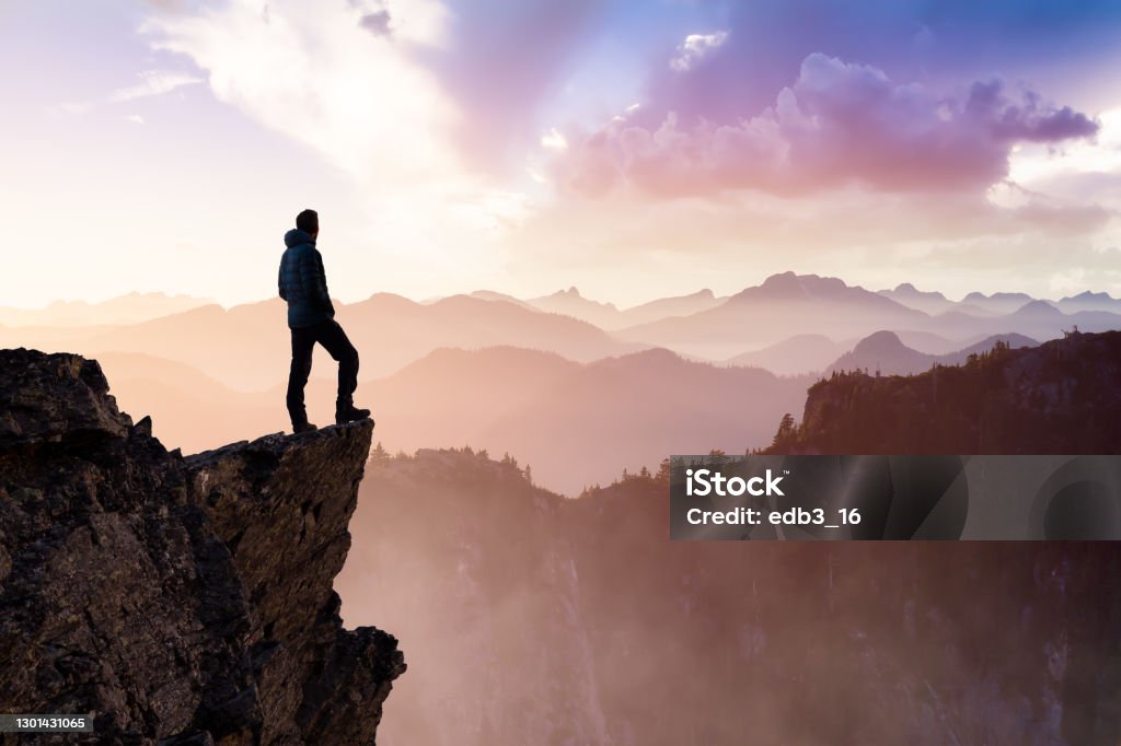 Man Hiker on top of a Mountain Peak. Composite. Adventurous Man Hiker With Hands Up on top of a Steep Rocky Cliff. Sunset or Sunrise. Landscape Taken from British Columbia, Canada. Concept: Adventure, Explore, Hike, Lifestyle Courage Stock Photo