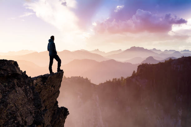 Man Hiker on top of a Mountain Peak. Composite. Adventurous Man Hiker With Hands Up on top of a Steep Rocky Cliff. Sunset or Sunrise. Landscape Taken from British Columbia, Canada. Concept: Adventure, Explore, Hike, Lifestyle courage stock pictures, royalty-free photos & images