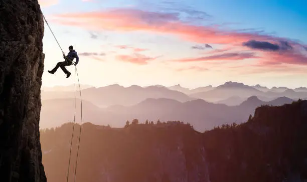 Adventurous Man Rappelling from Cliff. Beautiful aerial view of the mountains during a colorful and vibrant sunset or sunrise. Landscape in British Columbia, Canada. composite. Concept: Adventure