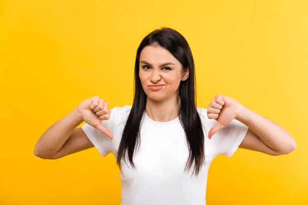 Dislike gesture. Displeased caucasian young woman wearing white basic t-shirt shows thumbs down gesture and negative grimace on her face, standing on isolated orange background