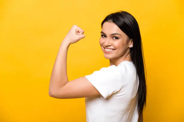 Photo of Young caucasian woman in a white t-shirt showing her strength by showing her hand while standing against an isolated orange background looks and smiles into the camera. Healthy and sporty lifestyle