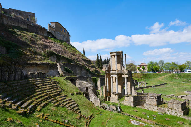 Remains of ancient arena and damaged building with columns located near hill with houses against cloudy sky