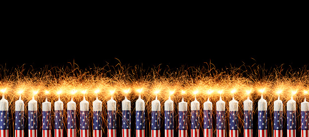 A long row of red, white and blue candles decorated in a patriotic American theme that are lit with sparkler flames against a black background.