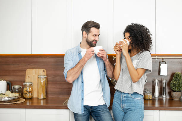 Interracial couple enjoying happy morning together Shy young African American woman and handsome man interracial couple enjoying the happy morning together, standing leaning on the modern kitchen counter and holding cup of coffee in their hands arabica coffee drink stock pictures, royalty-free photos & images