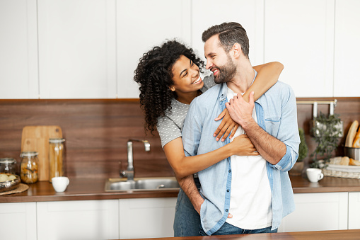 Close-up of a happy interracial couple standing in the kitchen, happy romantic owners of a new flat smiling and looking at each other, young African American woman hugging handsome man from behind