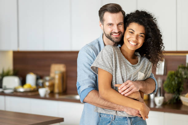 Two people hugging and joyfully looking at camera Smiling man hugging from behind charming African American woman, two people standing and joyfully looking at camera. Young international couple happily spending time in cozy modern kitchen at home. couple stock pictures, royalty-free photos & images