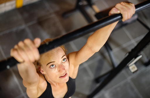 High angle of a determined-looking fit young woman doing chin-ups during a strength training workout in a gym