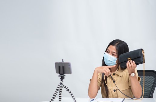 A young woman wearing a Covid-19 anti-virus mask, she is selling her purse online through her smartphone.