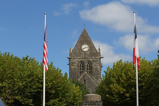 Sainte-Mère-Église, France - sep 1, 2014: a monument and the american and canadian flag with green trees in front and the church with the paratrooper and blue sky in the background