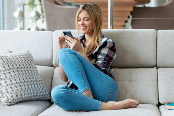 istock Pretty young smiling woman using mobile phone sitting on a couch at home. 1301467163