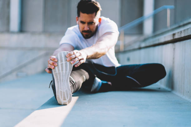 istock Blurred Caucasian sportsman doing stretching exercise during morning workout at urban setting, selective focus on strength male hands holding sneakers sole doing flexibility warm up with effort 1301474165