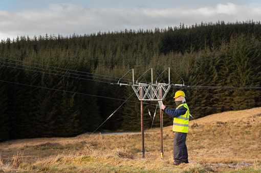 The rear view of a man wearing a high visibility waistcoat and hard hat using a digital tablet while standing close to high voltage power lines in rural Dumfries and Galloway south west Scotland