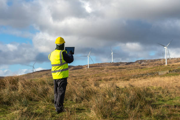 la vista posteriore di un uomo che guarda un tablet digitale mentre è fuori a lavorare in una posizione remota vicino alle turbine eoliche - galloway foto e immagini stock