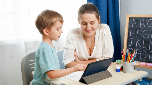 istock Little boy studying on digital tablet computer at home. Mother looking at er little son doing homework on tablet computer 1301487653