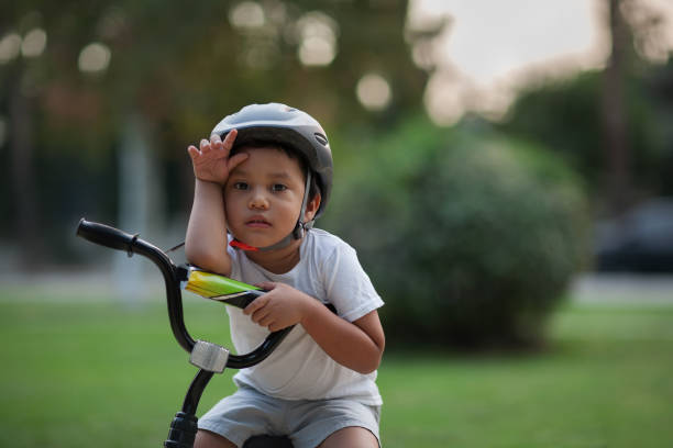 A little kid who looks exhausted or shows the difficulty in riding a bike for the first time by holding his hand to his forehead and looking unhappy. stock photo