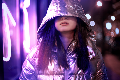 Young stylish asian woman wearing glossy jacket and hood covering eyes standing on the street at night in fluorescent violet neon lights playful