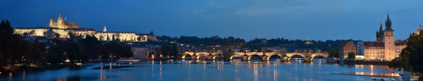istock Prague riverside at night. Panoramic image of illuminated Charles Bridge and riverside buildings with a clock tower 1301494840