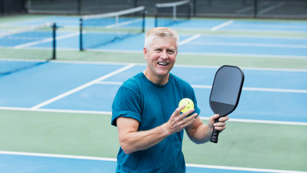 Senior man on pickleball court A senior man standing on a pickleball court, smiling at the camera, holding a paddle and ball. He is in his 60s. pickleball equipment stock pictures, royalty-free photos & images