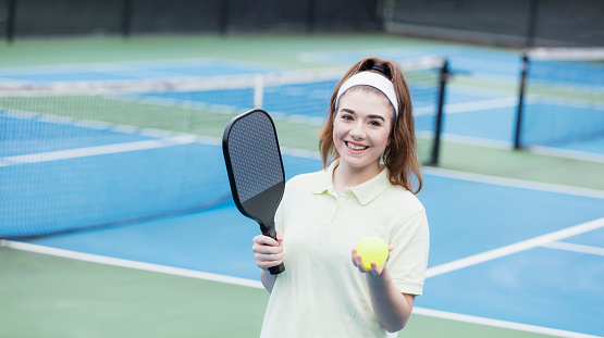 A young woman standing on a pickleball court, holding a paddle and ball. She is in her 20s.