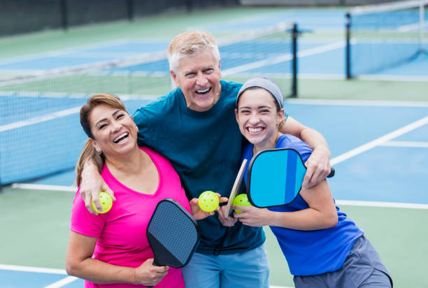 Couple and adult daughter playing pickleball An interracial family playing pickleball together. The mother is a mature Hispanic woman in her 50s. Father is a senior in his 60s. Their adult daughter, in her 20s, is mixed race Hispanic and Caucasian. They are smiling and laughing, looking at the camera. pickleball stock pictures, royalty-free photos & images