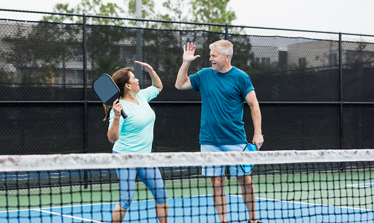 A couple playing pickleball together. They are a doubles team. The Hispanic woman, in her 50s, has just hit a winning shot and her partner, a senior man in his 60s, is giving her a high-five