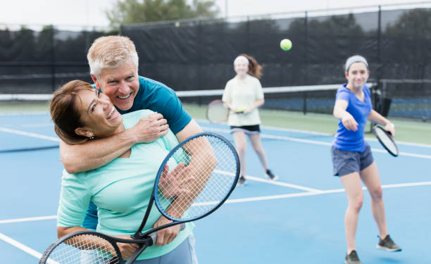 Couple playing tennis, with family, hugging An interracial family playing tennis together. The mother is a mature Hispanic woman in her 50s. Father is a senior in his 60s. Their adult daughter, in her 20s, is mixed race Hispanic and Caucasian. The focus is on the parents standing in the foreground, smiling at the camera, hugging.. tennis senior adult adult mature adult stock pictures, royalty-free photos & images