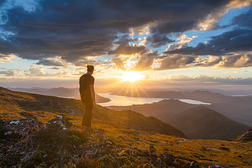 Man standing on cliff watching sunset over golden mountain range, Tasmania, Australia