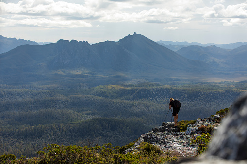 Young male photographer standing on edge of cliff taking photos of mountain in background in Tasmania, Australia
