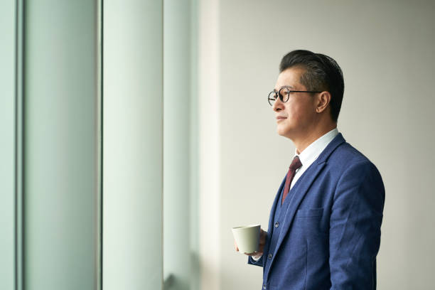 asian business manager standing in front of window thinking - business business person ceo coffee imagens e fotografias de stock