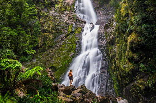 Young man standing below waterfall, with green moss and dense rainforest environment, Tasmania, Australia
