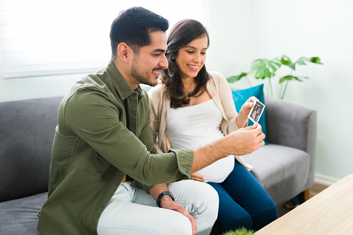 Smiling pregnant woman sitting on the sofa and  showing her cheerful husband a print ultrasound of the baby she carries in her belly