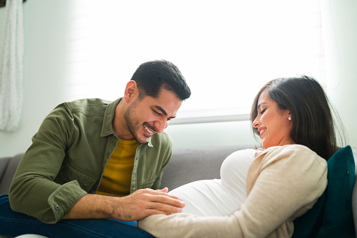 Good-looking latin man looking and touching his pregnant wife's belly. Smiling caucasian wife sharing her dreams and thoughts about their future baby with her husband