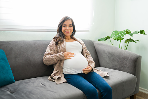 Beautiful pregnant woman in her 30s wearing casual clothes and sitting on her home living room. Happy expectant mother resting on the couch