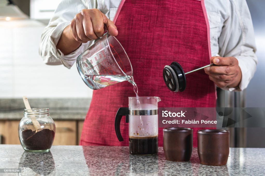 Horizontal shot of a man in a red apron placing boiling water in a French press to prepare coffee Horizontal shot of a man in a red apron placing boiling water in a French press to prepare coffee. Barista whose face is not visible. Selective focus Water Stock Photo