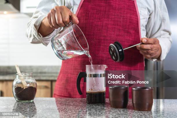 Horizontal Shot Of A Man In A Red Apron Placing Boiling Water In A French Press To Prepare Coffee Stock Photo - Download Image Now