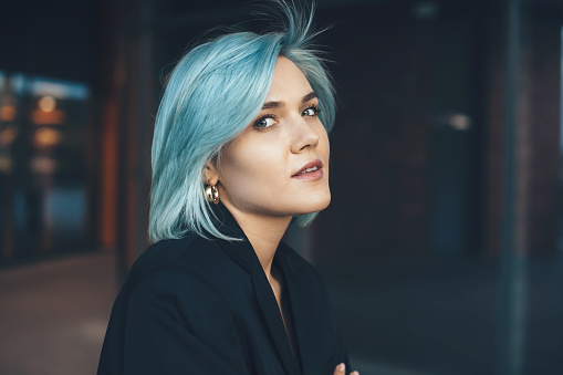 Young charming lady with blue hair posing outside and looking at camera with a building on background