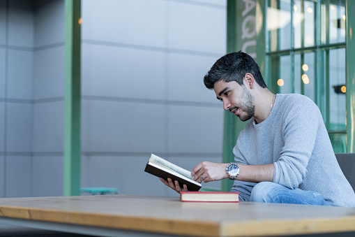 Latino ethnic man between 20-30 years old is sitting smiling reading a book in a shopping center