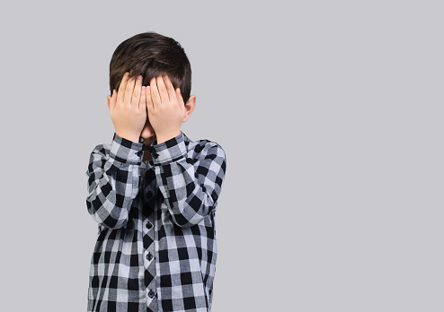 Unhappy schoolboy, 6-year-old boy covered his face with his hands on studio gray isolated background.Resentful boy