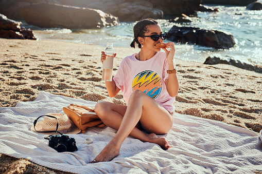 Shot of a woman holding a water bottle and a donut while sitting on the beach