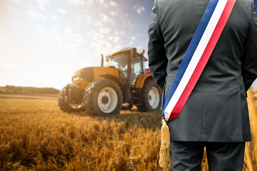 Male farmer with short brown hair,wearing a gray pullover and black vest walking in the direction to his tractor,standing next to his agricultural field in the evening with lights turned on,focus on foreground,low angle view