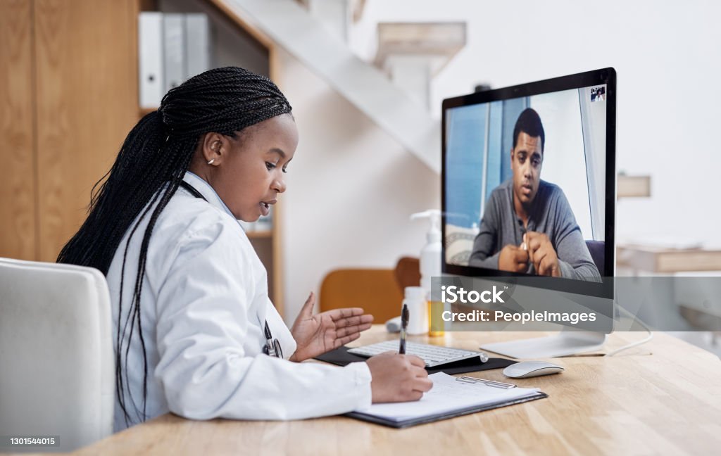 Taking notes during her virtual consult Shot of a young doctor writing notes during a video call with a patient on a computer Telemedicine Stock Photo