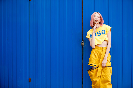 Street Fashion Woman with dying Pink Hair color wear yellow chic dress. Young Girl poses on Strong blue colorful wall art as summer trend style. copy space
