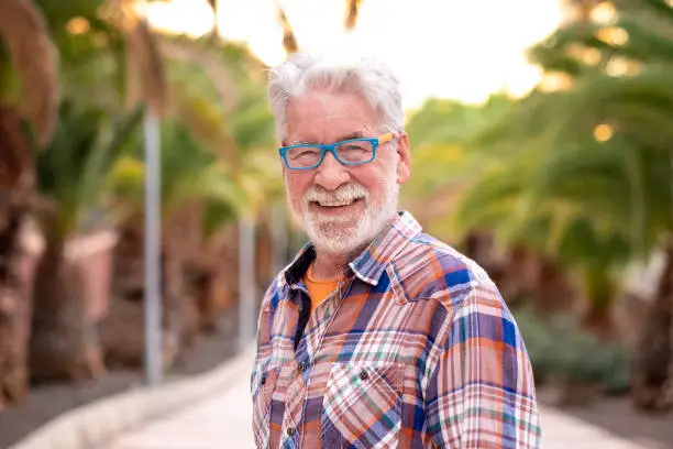 Portrait of attractive senior man white-hair and beard looking at camera. Standing outdoor in a public park at sunset. A smiling and relaxed people with checkered shirt