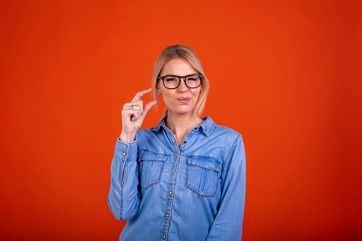 Positive woman in denim dress showing a little bit gesture and looking with disappointment, measuring centimeter, inch size. indoor studio shot isolated on orange background