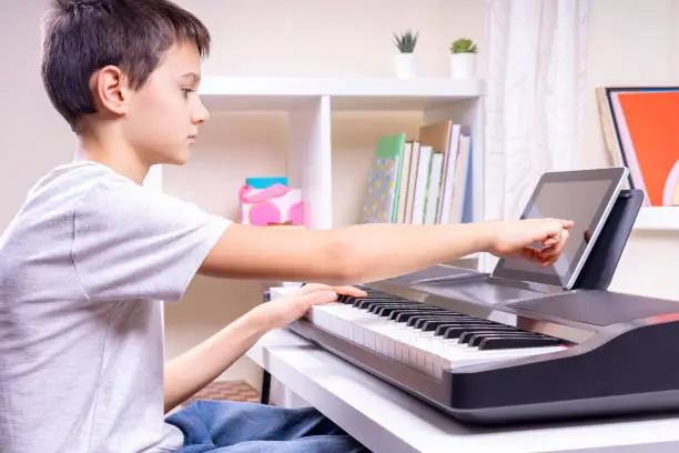 Photo of Boy learning music online. Child watching video tutorial at tablet computer and playing digital piano at home