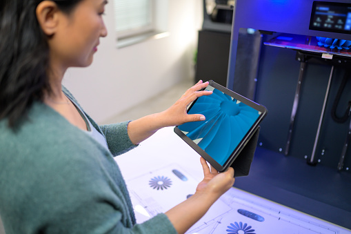 Female engineer holding a digital tablet and checking the propeller design while standing next to a 3D printer.