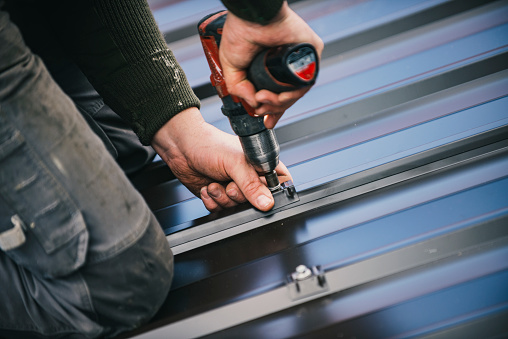 Building contractor is installing metal roofing sheets on the rooftop of the house using electric screwdriver.